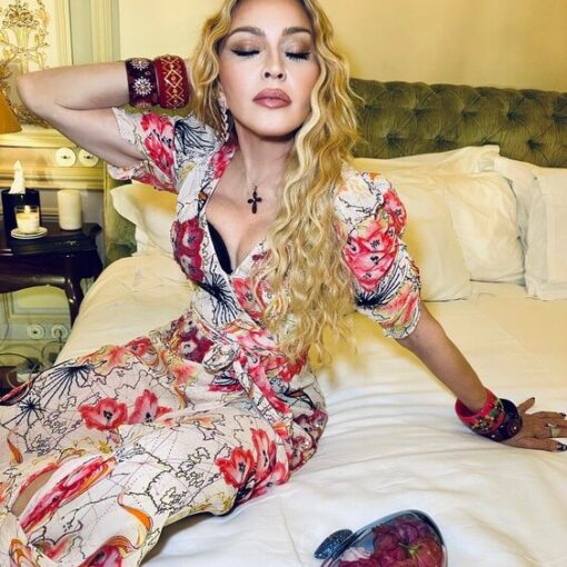 Madonna in a floral dress with low cut cleavage and black bra peaking out in tasteful photo sitting on a bed for her 65th Birthday celebration.