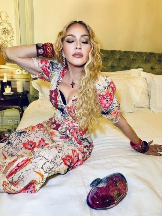 Madonna in a floral dress with low cut cleavage and black bra peaking out in tasteful photo sitting on a bed for her 65th Birthday celebration.